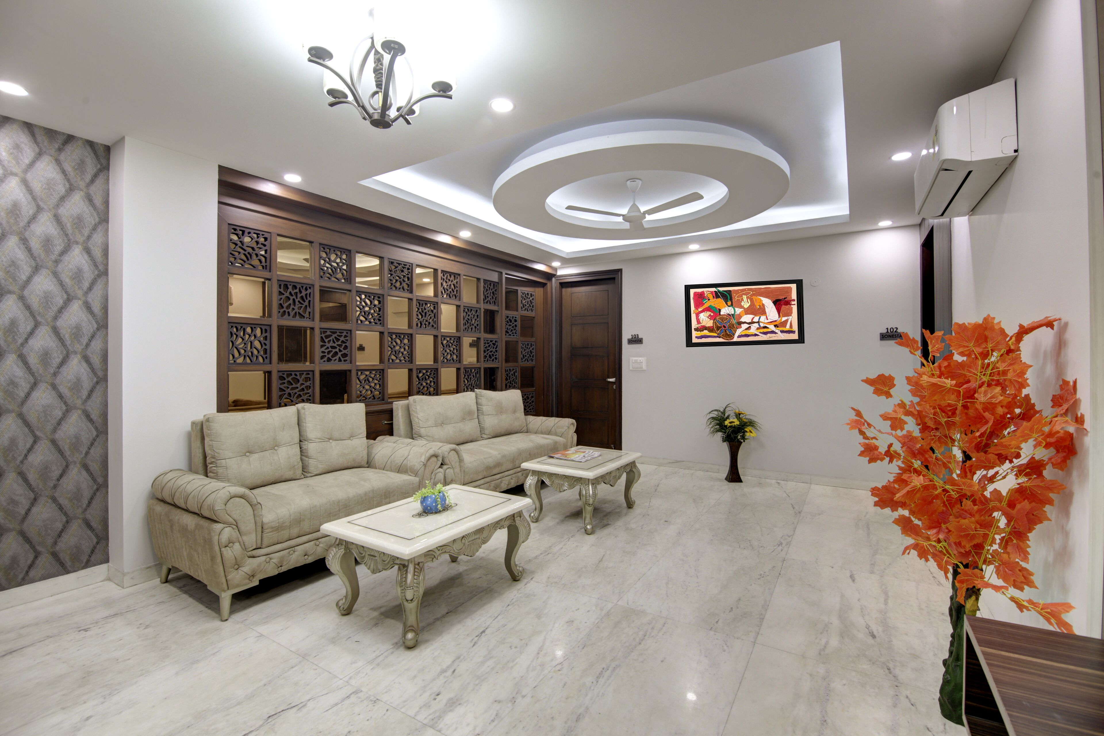 A modern living room with a sofa set, two coffee tables, a decorative wall panel, a ceiling light fixture, and a potted plant.