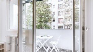 Une pièce avec un radiateur blanc, une table blanche et deux chaises, et une vue sur un immeuble doté de balcons et d’arbres, visible à travers une porte vitrée.