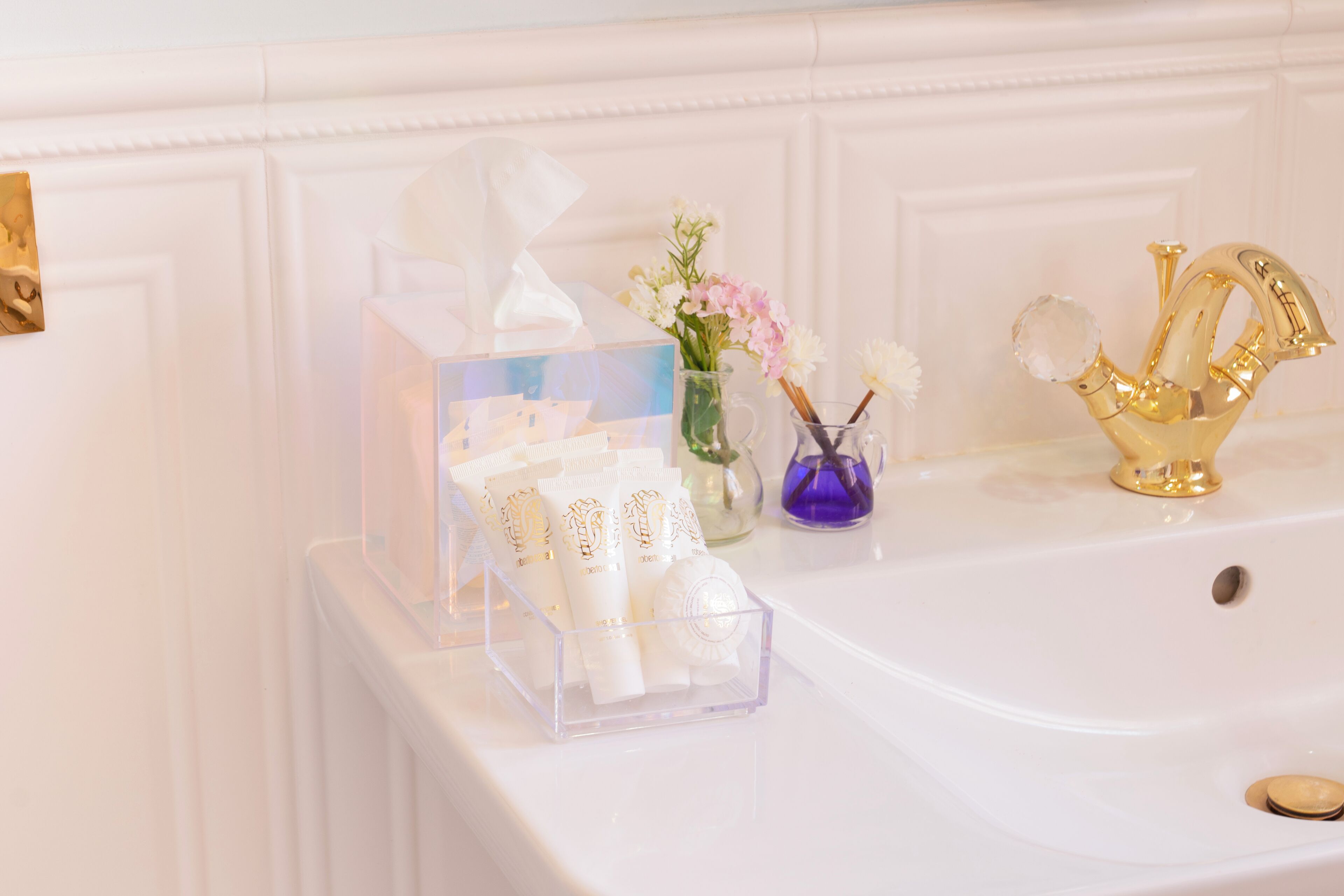 A bathroom vanity with a clear tissue box, a vase with flowers, and a golden faucet.