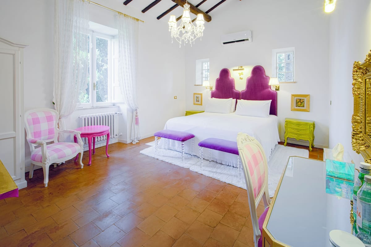 Een slaapkamer met een bed, twee stoelen, een tafel, een kroonluchter en een raam.