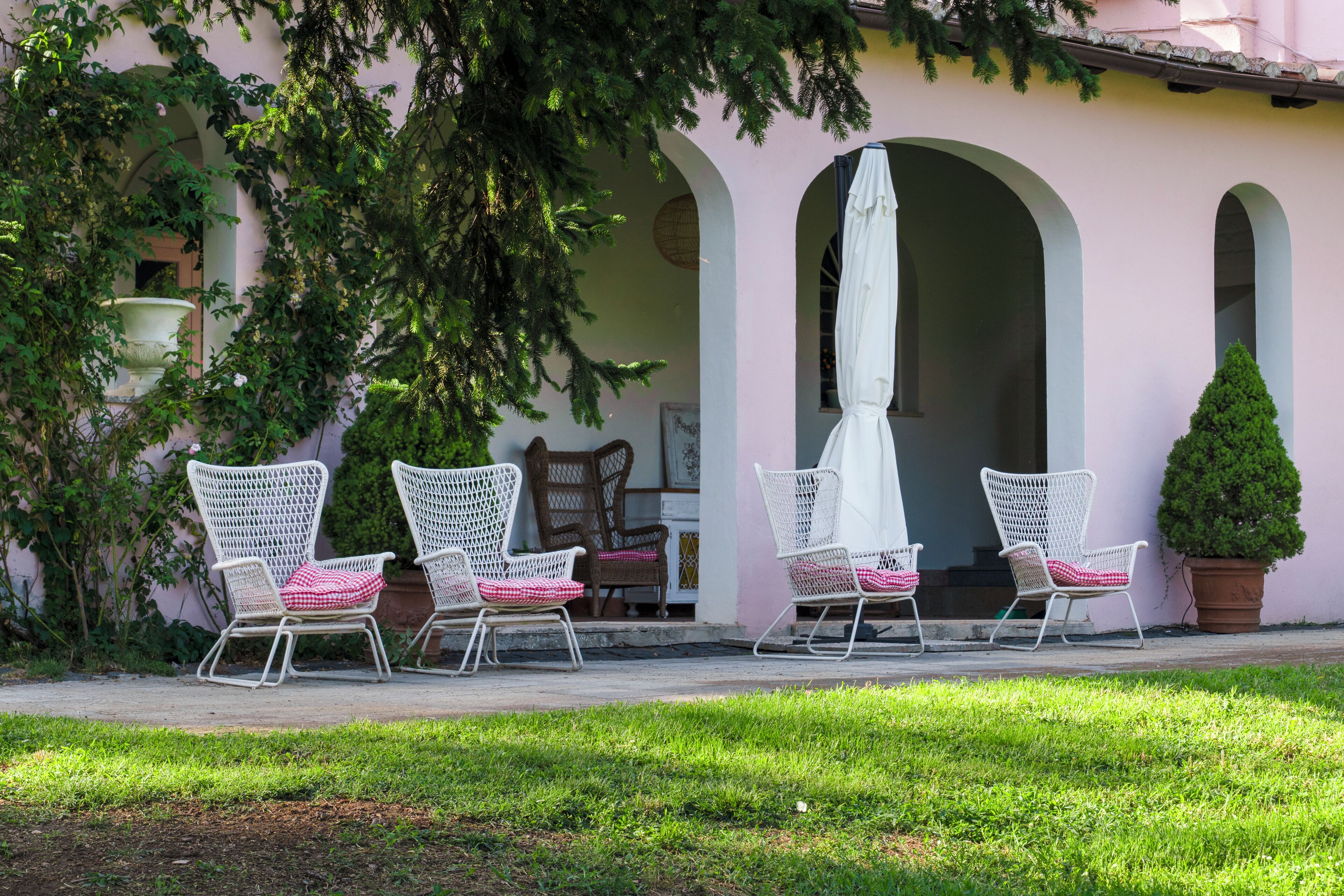 A patio with white wicker chairs and a white umbrella, surrounded by greenery and a pink building with arches.