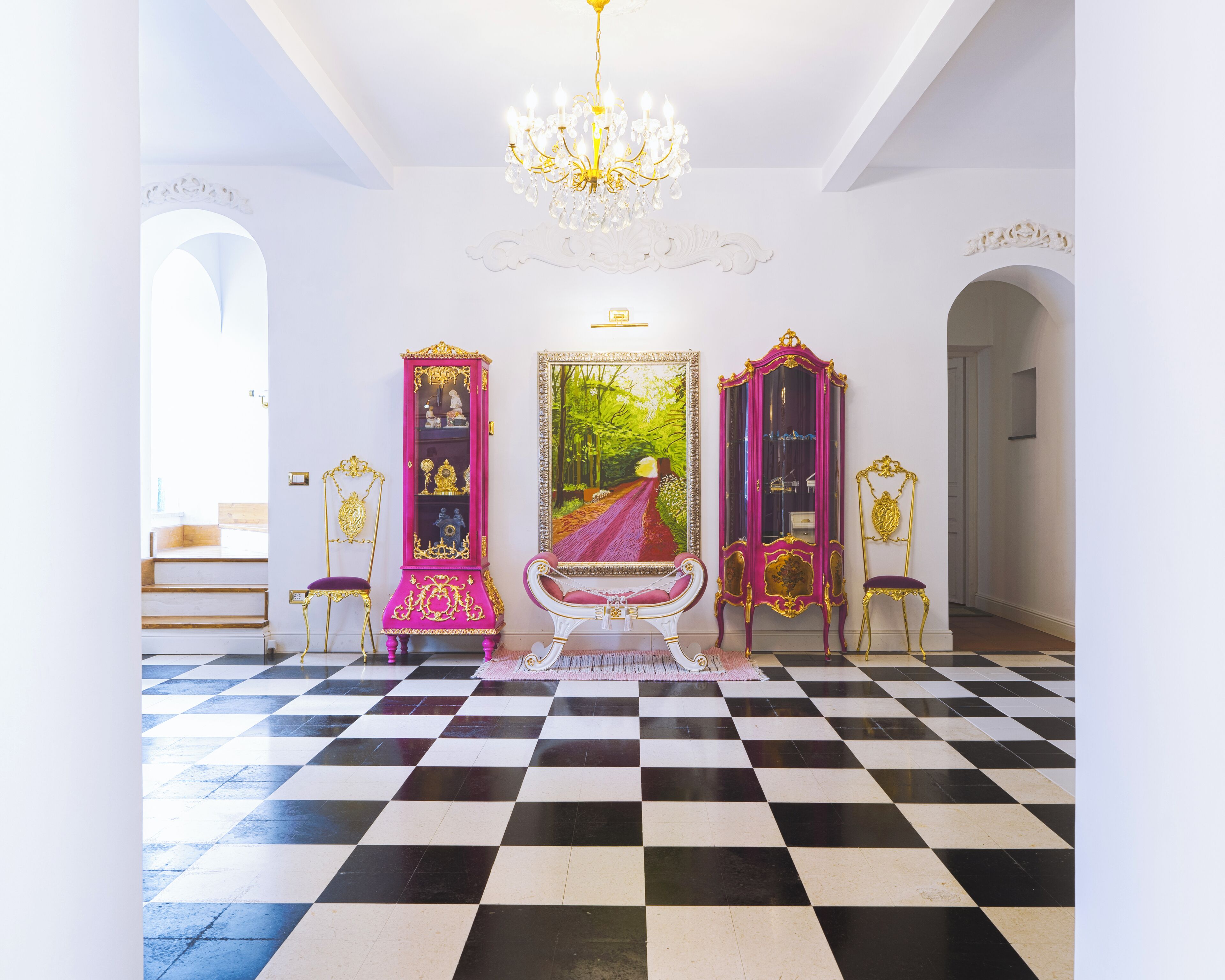 A grand hallway with a black and white checkered floor, ornate gold and pink furniture, and a large mirror with a forest painting.