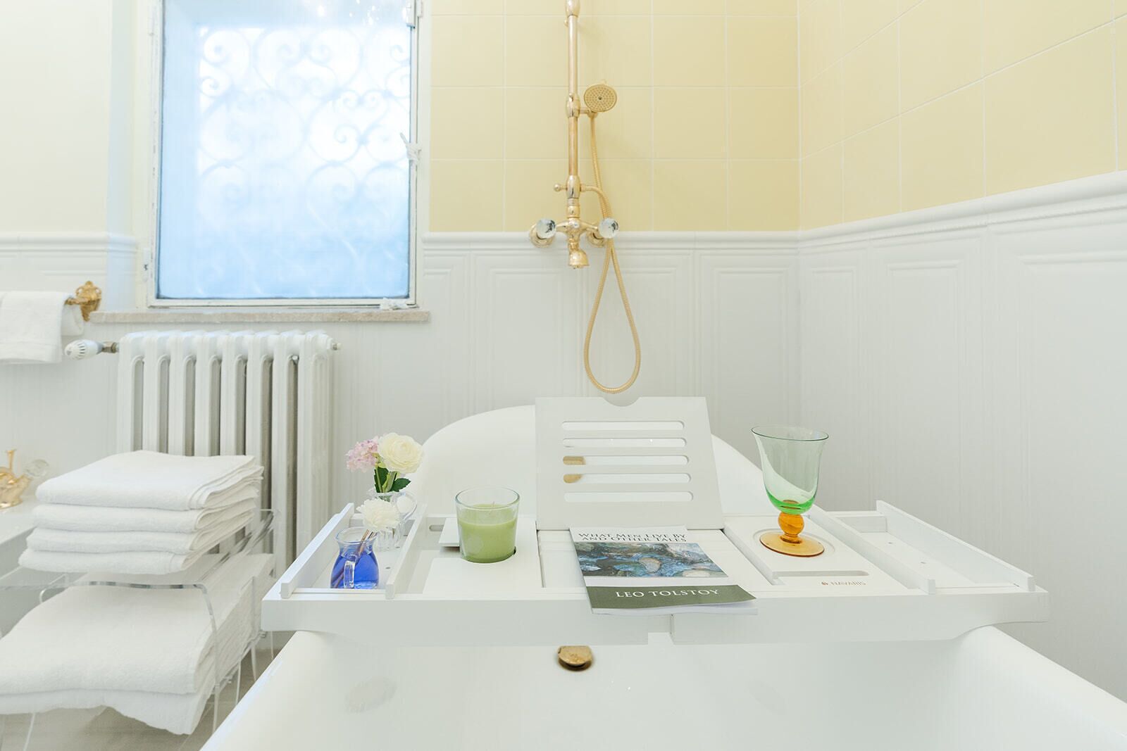 Un baño con una bañera blanca, un vaso de líquido verde, un libro titulado 'Leo Tolstoy' y una ventana con vistas al cielo.