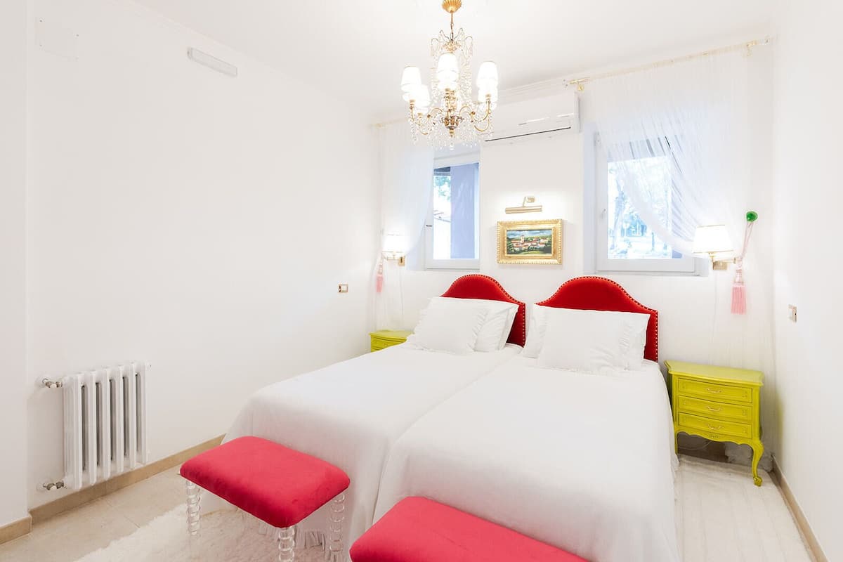 Een tweepersoonsbed met rood hoofdeinde en voeteinde, witte beddengoed, een geel nachtkastje met lamp, en een radiator.
