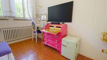 Ett rum med en rosa byrå, ett vitt element, ett litet vitt kylskåp, en tv på väggen och ett fönster med gardiner.