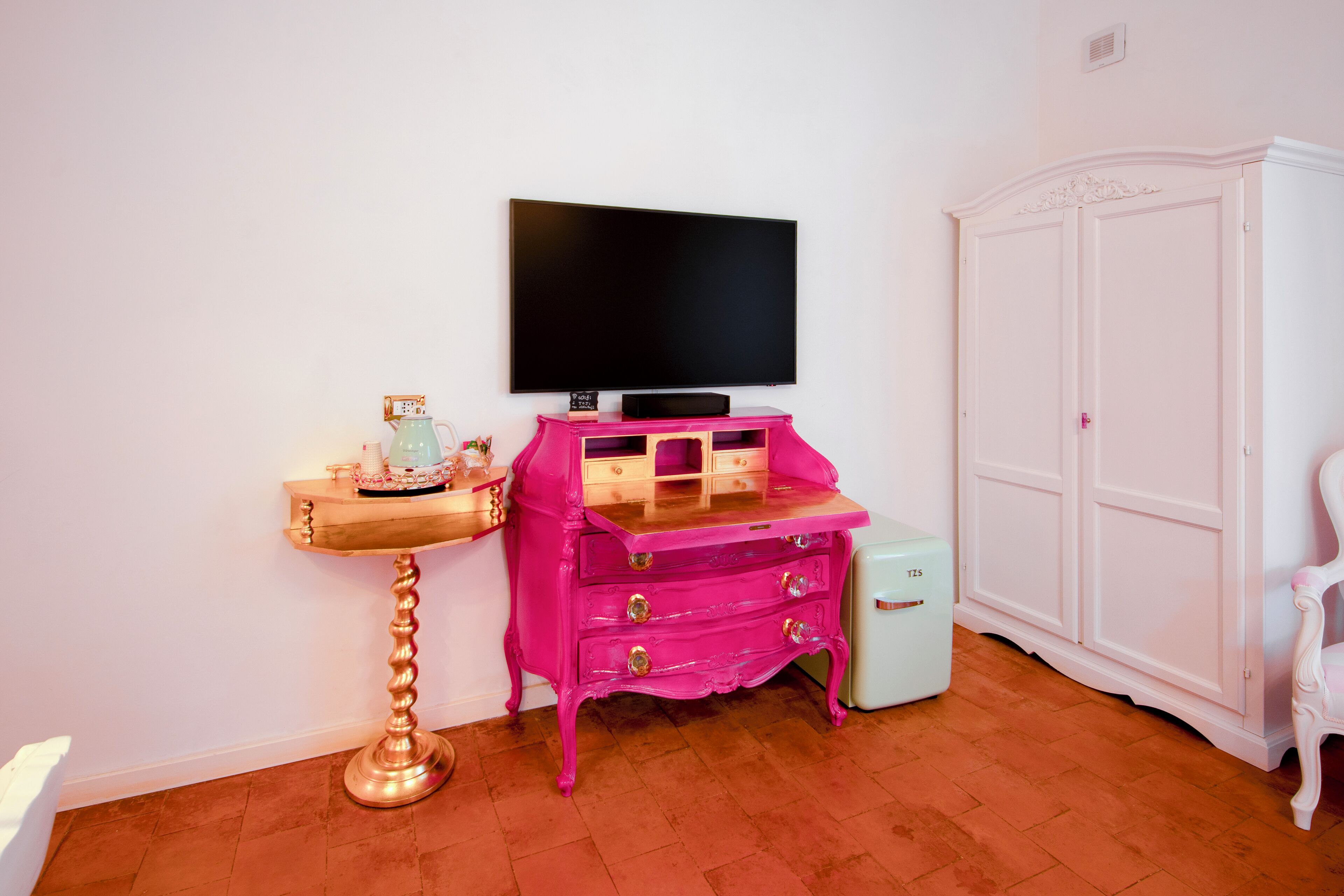 A room with a flat-screen TV mounted on the wall, a pink dresser with drawers, a small white cabinet, and a white wardrobe.
