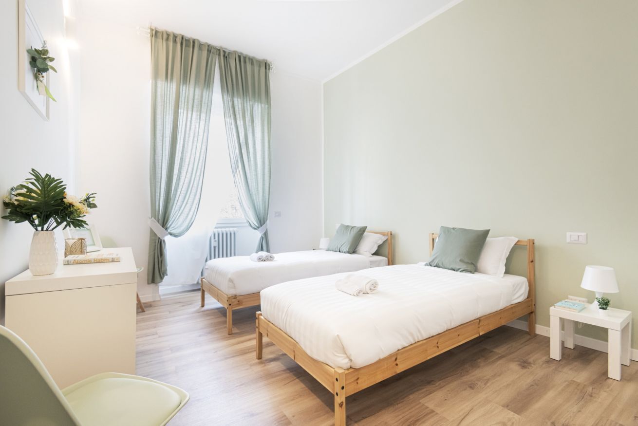 A bedroom with two single beds, a bedside table with a lamp, a chair, and a window with curtains.