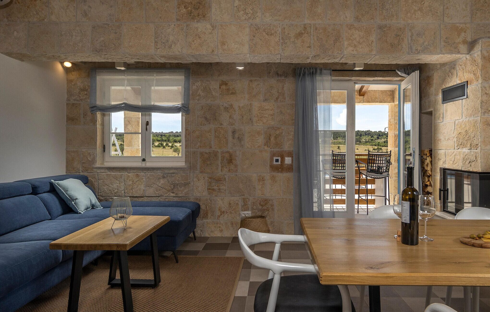 Una sala de estar acogedora con un sofá azul, una mesa de centro de madera y una zona de comedor con mesa y sillas de madera. Se observa una chimenea a la derecha, y una ventana ofrece vistas al exterior.