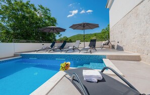 Un espace piscine aménagé avec des chaises longues et des parasols, une piscine carrelée et une vue sur la verdure et les collines.