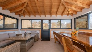 A dining area with a wooden table and benches, a wicker sofa, and a view through large windows. - Nice home in Dobrince (Dobrince)