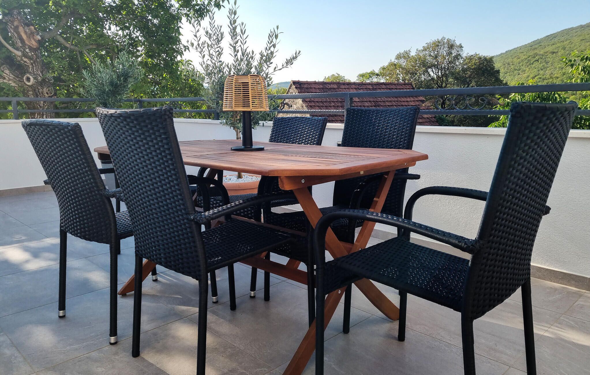 Een ronde houten tafel met zwarte rieten stoelen, een geweven mand en een metalen hekwerk op de achtergrond.
