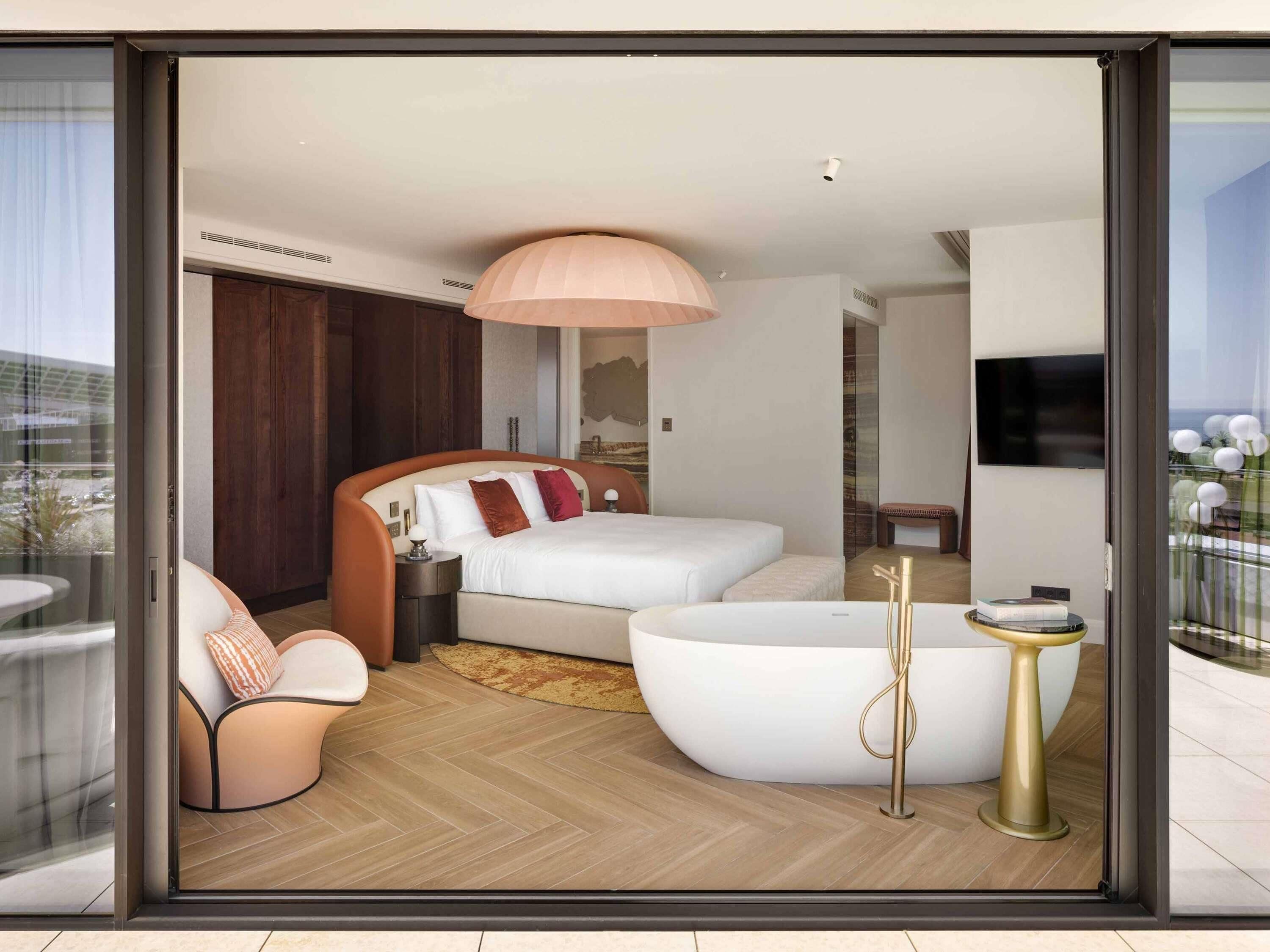 A modern hotel room with a large bed, a sitting area with a chair, a bathtub, and a television.