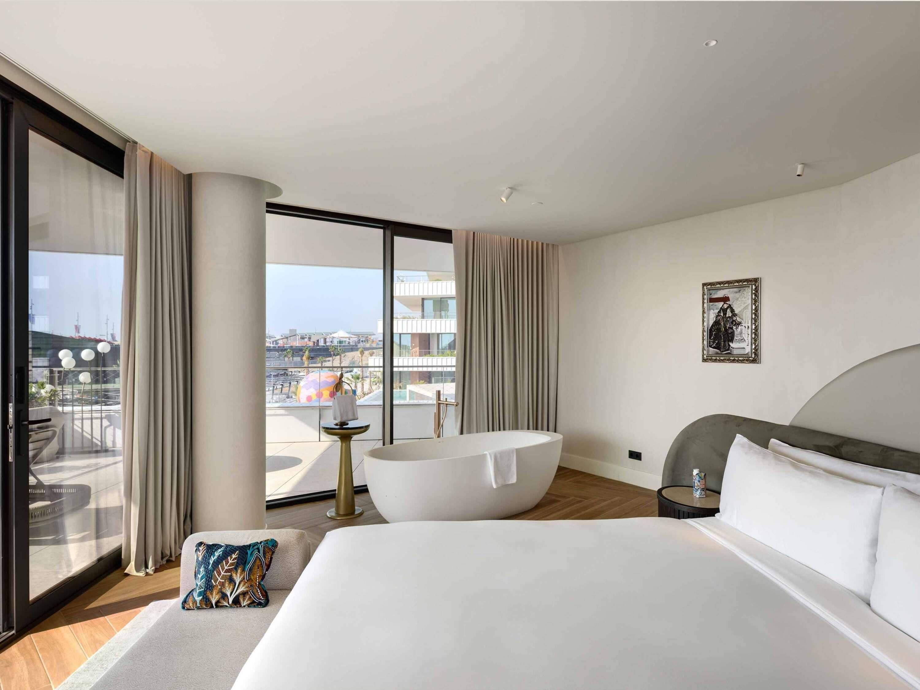 Habitación de hotel moderna con una cama grande, una bañera y un balcón con vistas a la ciudad.