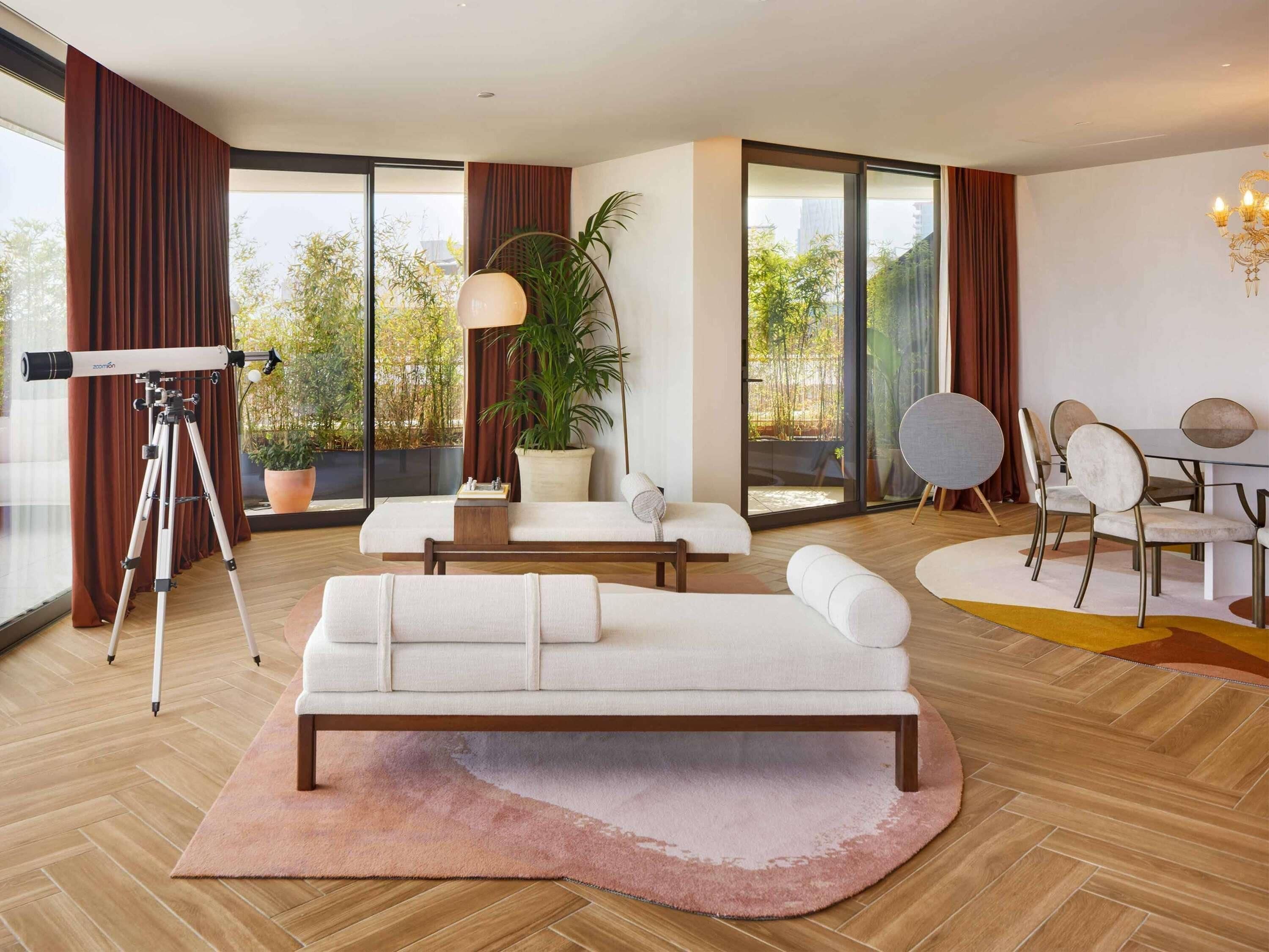 A modern living room with a sofa, a dining table with chairs, a telescope, and a large window with curtains.