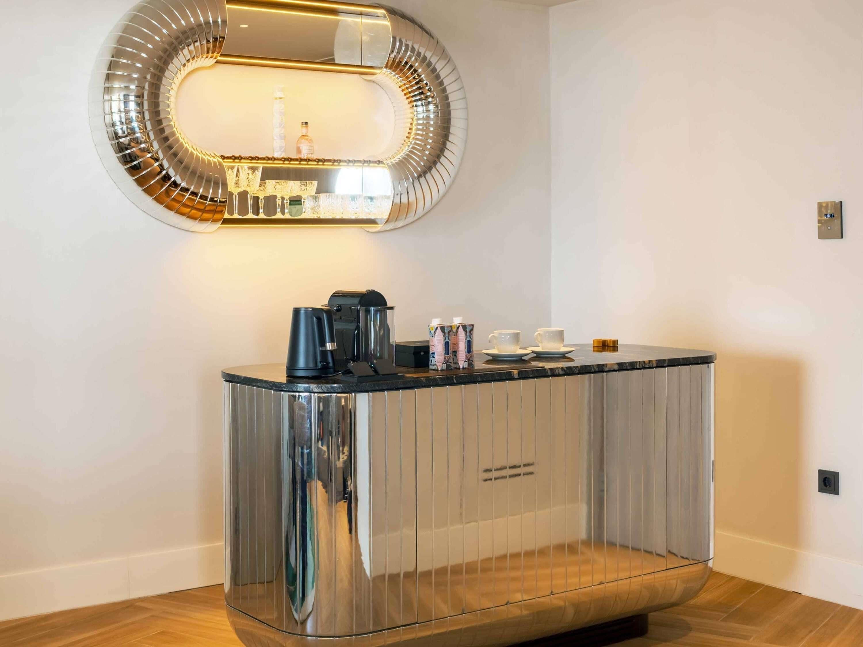A modern, metallic-framed mirror above a bar area with a coffee machine, cups, and condiments.