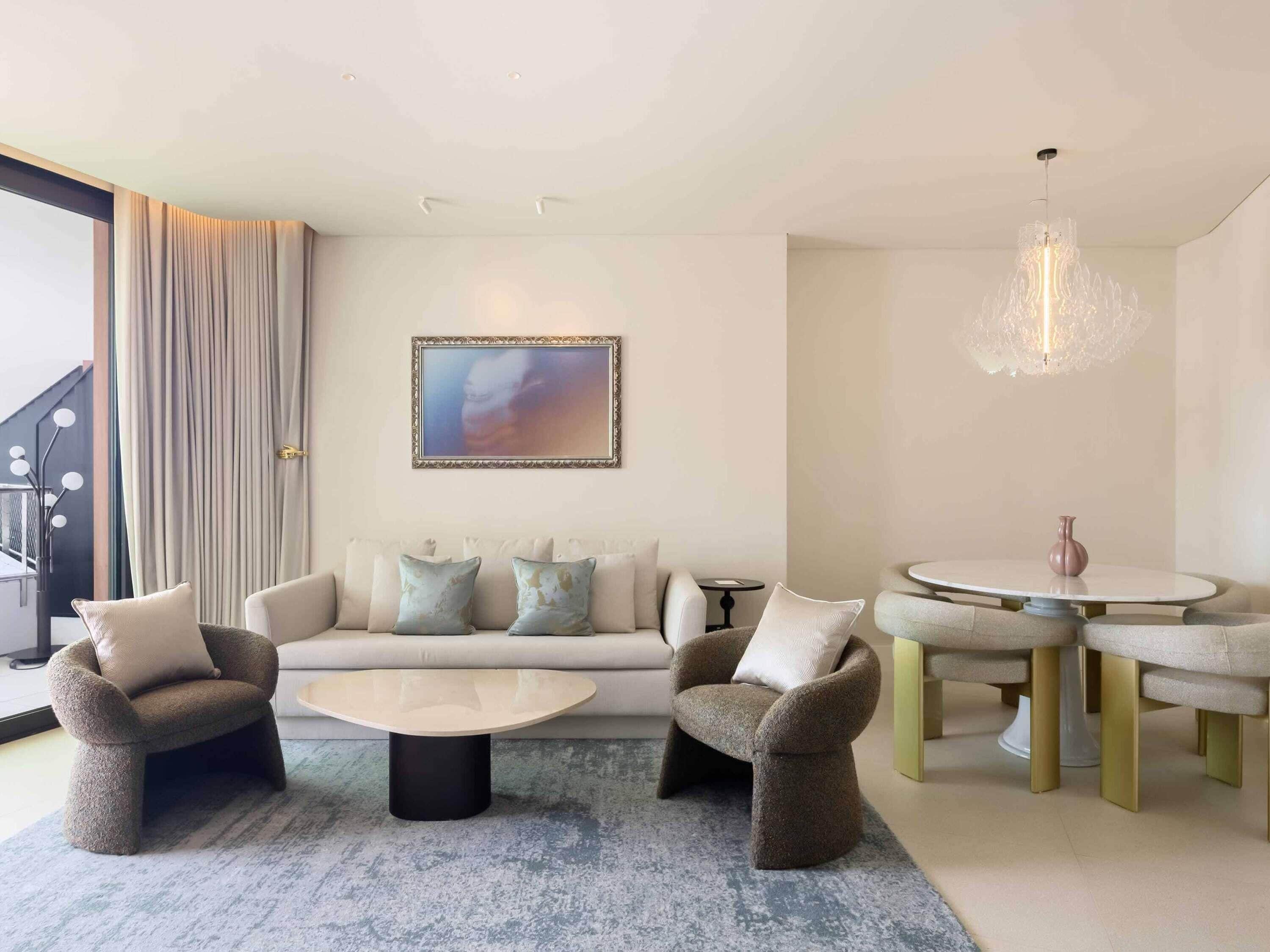 Een moderne woonkamer met een bank, twee fauteuils, een salontafel, een eettafel met stoelen, een kroonluchter en een groot raam met gordijnen.