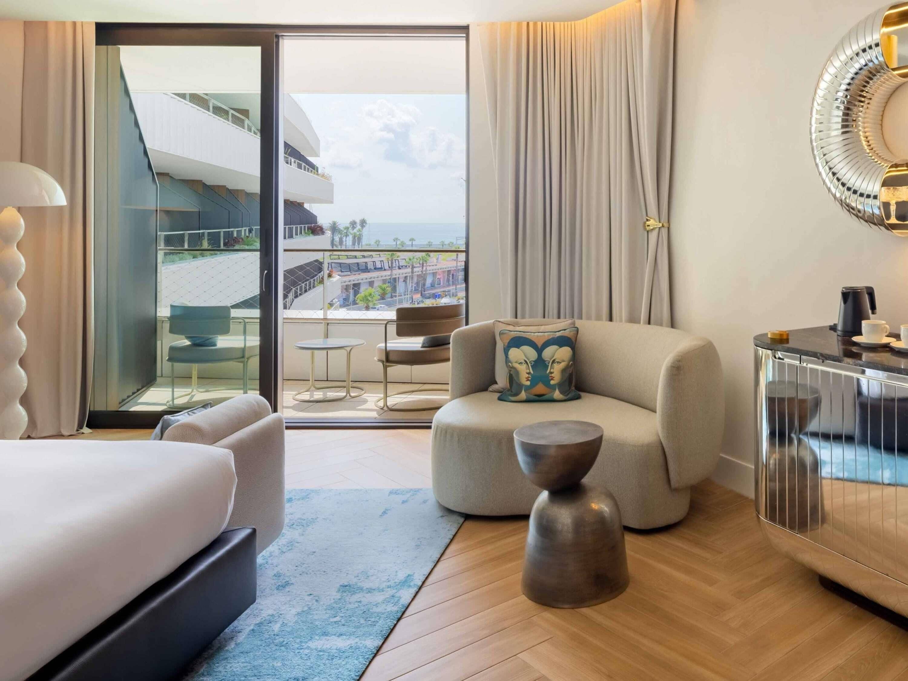 Habitación de hotel con cama, sofá, mesa de centro y vistas a un edificio y palmeras.