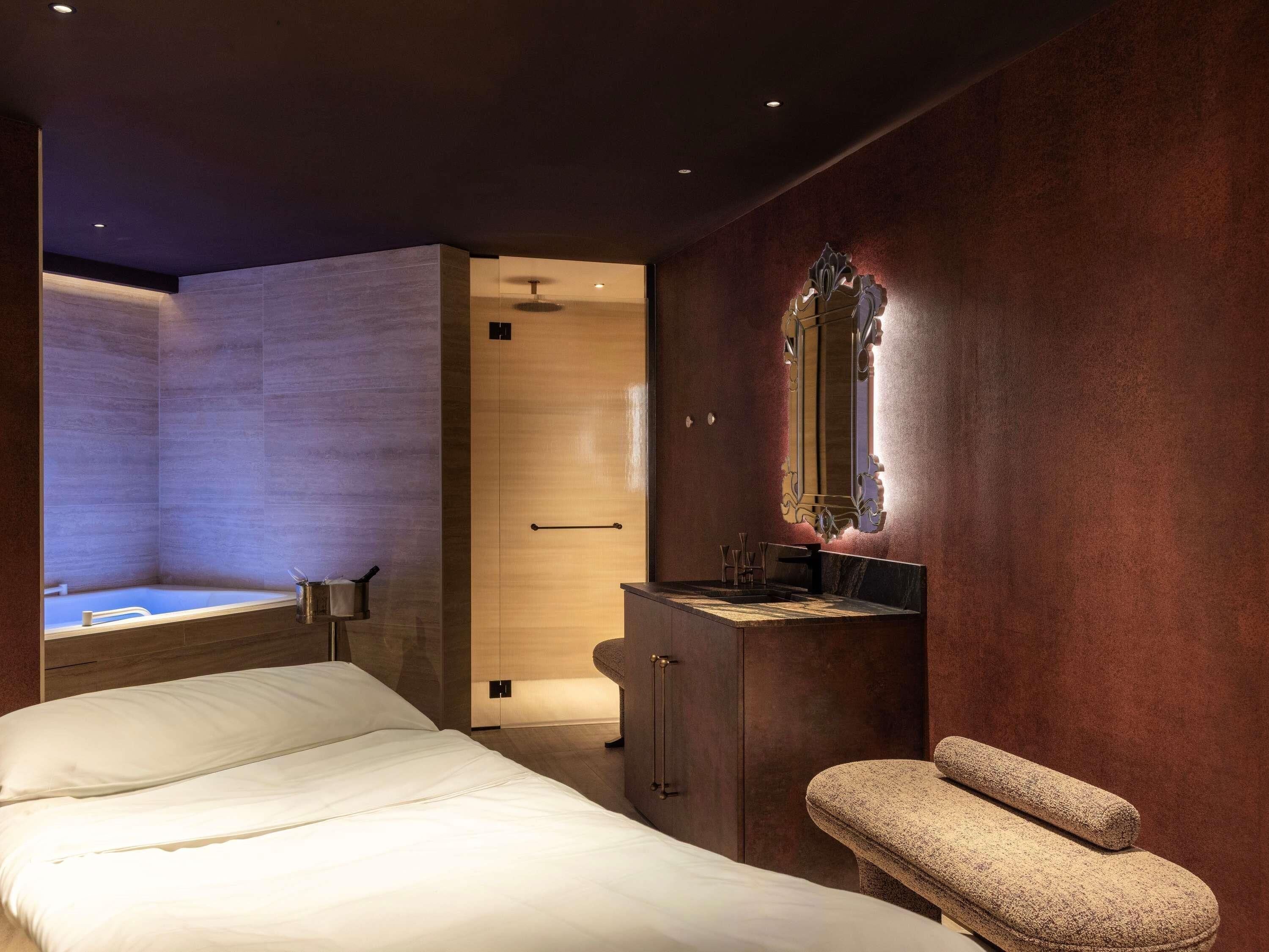 Una habitación de hotel moderna con una cama, un baño con bañera, un tocador con espejo y una silla de masaje.