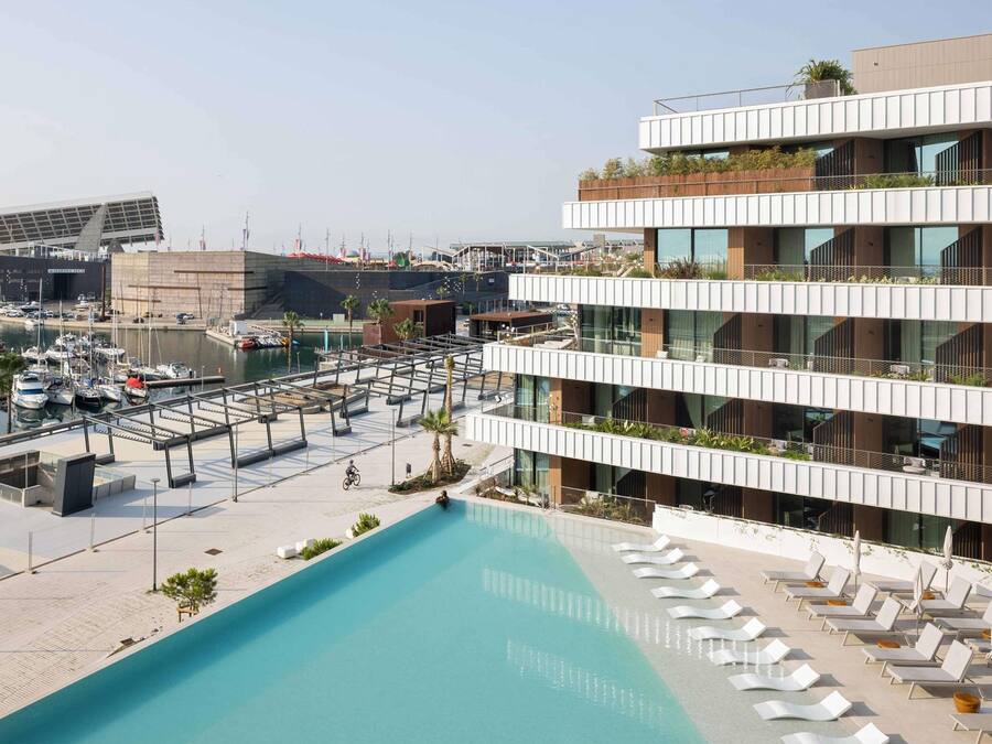 Um edifício moderno com piscina no terraço, várias varandas e um porto de iates ao fundo.