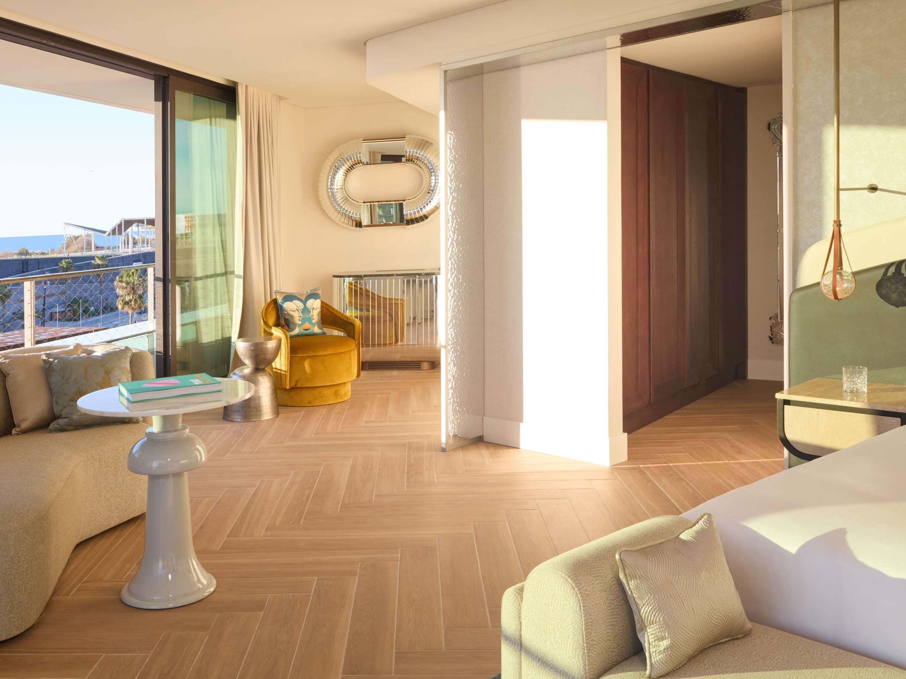 Quarto de hotel moderno com uma cama grande, um sofá, uma mesinha e uma cadeira. Possui uma varanda com vista para o mar.