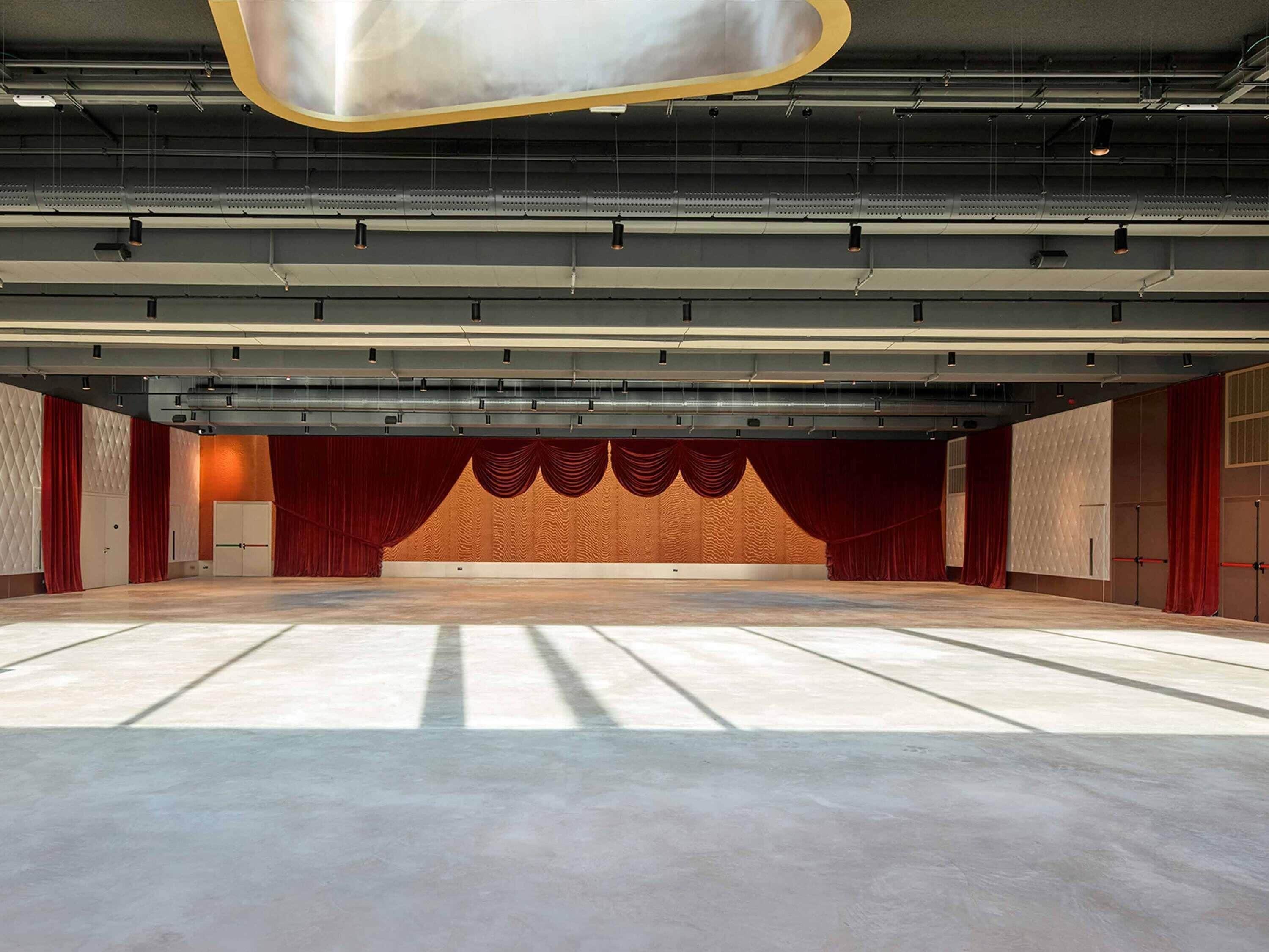 Um amplo auditório vazio com palco, cortinas vermelhas e um design de teto moderno.
