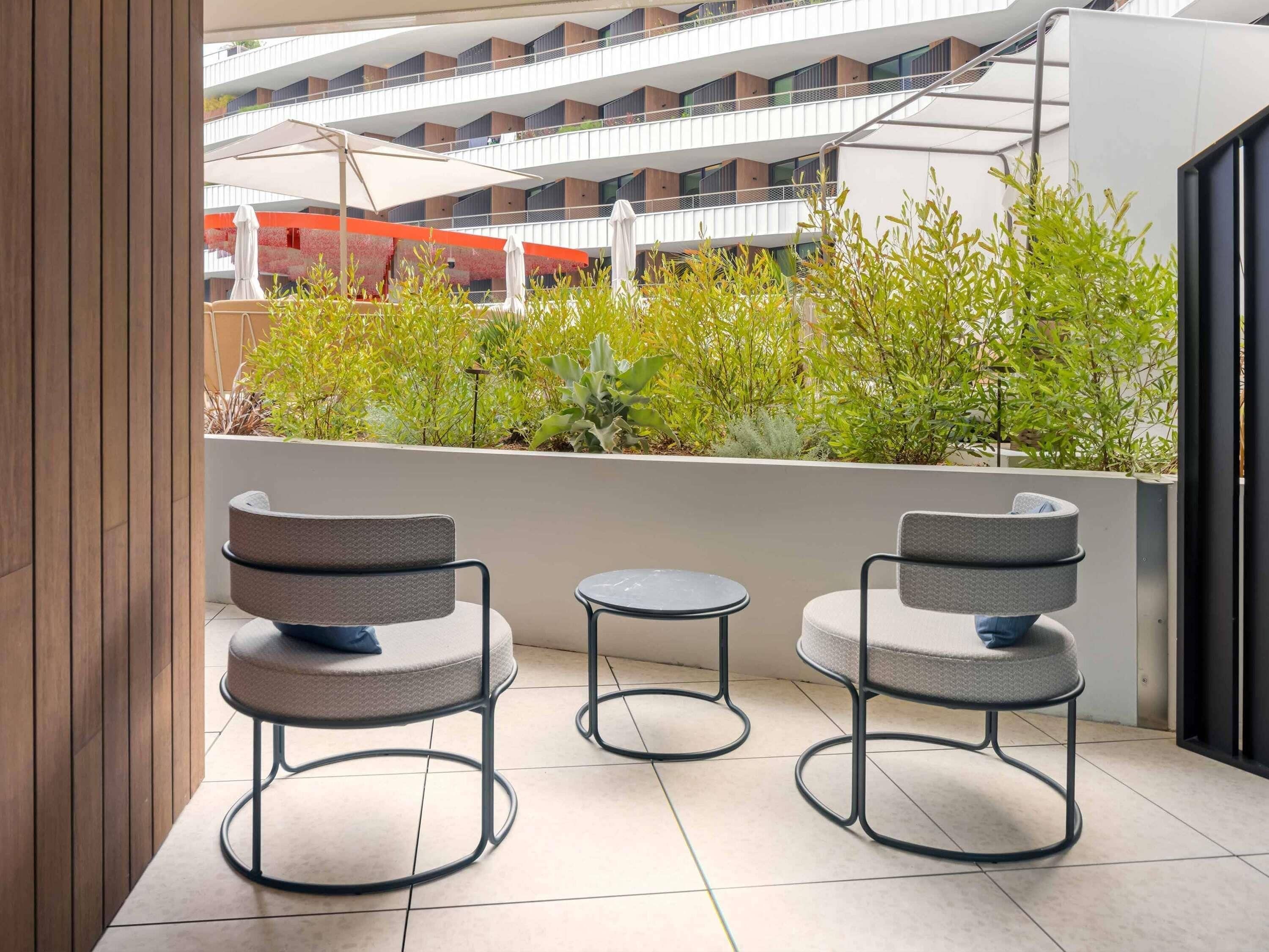 A balcony with two chairs and a small table, surrounded by greenery and overlooking a modern building.
