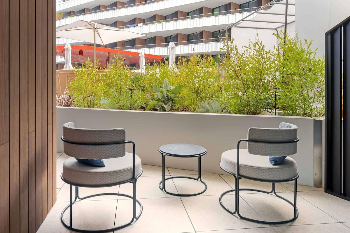 A balcony with two chairs and a small table, surrounded by greenery and overlooking a modern building.