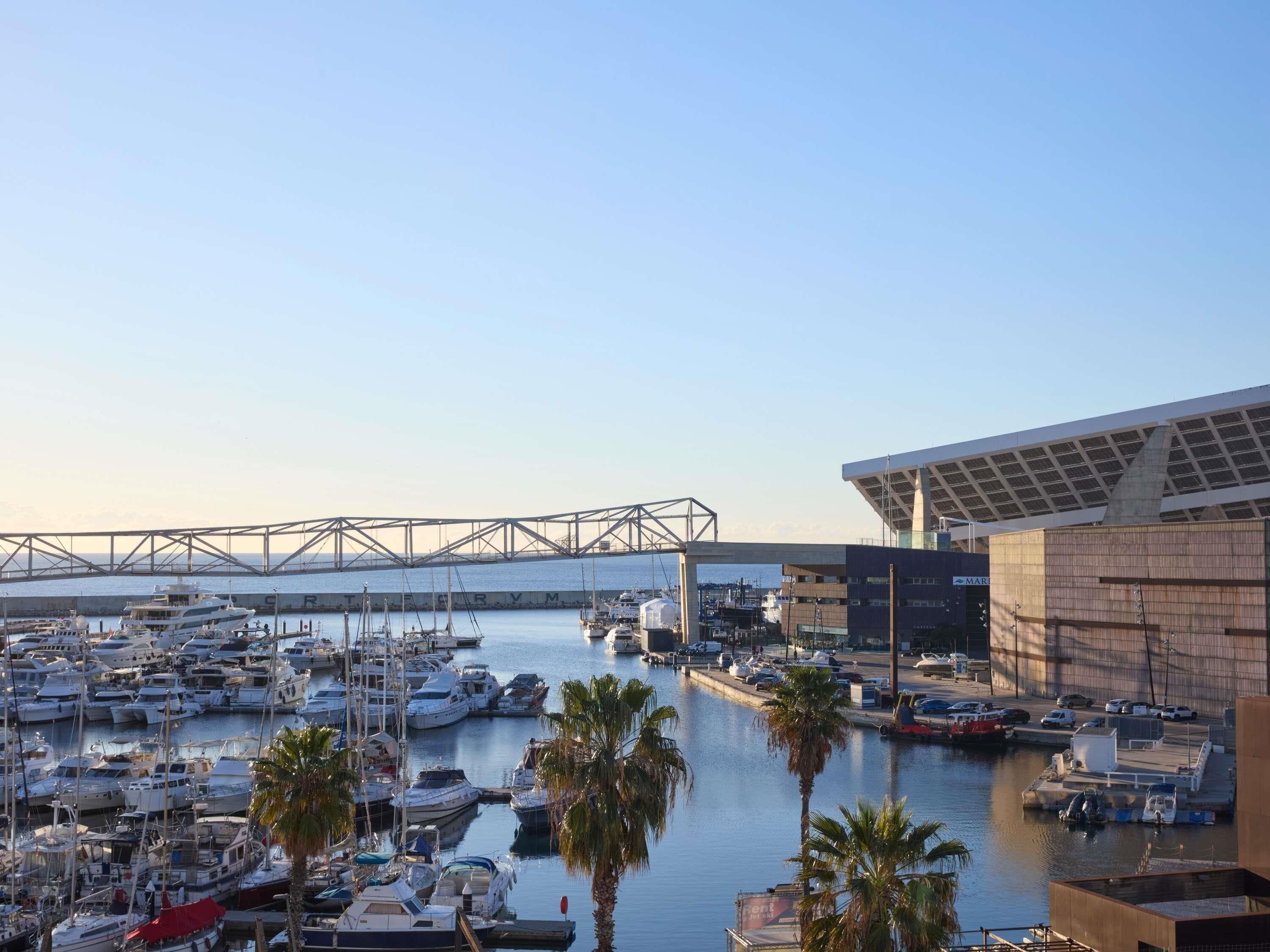A marina with numerous boats docked, a bridge in the background, and a modern building with a large roof structure.