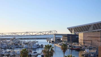 A marina with numerous boats docked, a bridge in the background, and a modern building with a large roof structure.