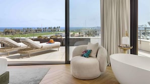 Een moderne hotelkamer met een ruim balkon, een ronde fauteuil, een badkuip en uitzicht op zee en de skyline.