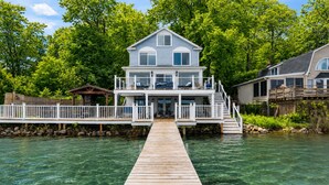 A two-story house with a dock leading to a private pier, surrounded by lush greenery. - Lakefront | Hot Tub | 10 Guest | Dock + Kayaks (Seneca Falls)