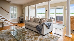 A spacious living room with a beige sofa, a glass coffee table, and a staircase leading to an upper level. - Lakefront | Hot Tub | 10 Guest | Dock + Kayaks (Seneca Falls)