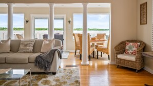 A spacious living room with a large window overlooking a body of water, a comfortable sofa, wicker chairs, and a dining area. - Lakefront | Hot Tub | 10 Guest | Dock + Kayaks (Seneca Falls)
