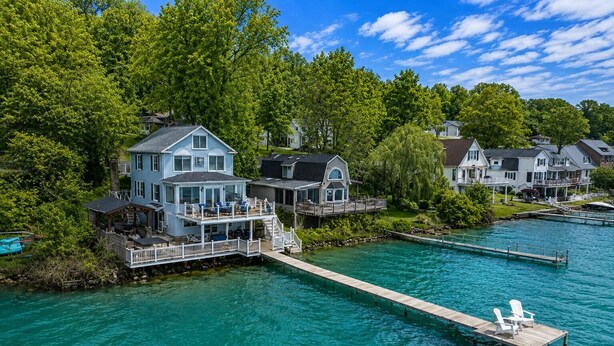 A waterfront property with a dock, a house with a balcony, and a second house in the background. - Lakefront | Hot Tub | 10 Guest | Dock + Kayaks (Seneca Falls)
