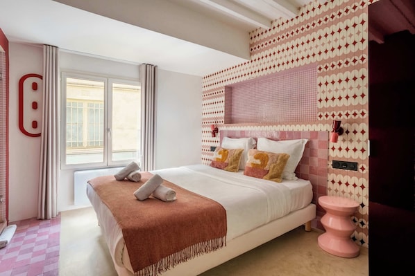 A bedroom with a bed, a window, a pink stool, and patterned wall tiles.