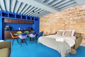 Quarto com cama, mesa de jantar com cadeiras coloridas, cozinha com armários e uma parede de pedra.