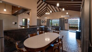 A spacious dining area with a marble countertop and wooden chairs, a high ceiling with exposed beams, and a view of the outdoors through large windows.