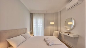 A hotel room with a large bed, a bedside table, a lamp, a mirror, and a built-in air conditioning unit. - Éclair: Lovely 2BD apt perfect for families (Thessaloniki)