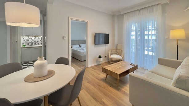 A modern, well-lit living room with a dining area, a sofa, a coffee table, and a TV mounted on the wall. - Éclair: Lovely 2BD apt perfect for families (Thessaloniki)