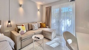 Ein modernes Wohnzimmer mit einer Eckcouch, einem kleinen runden Tisch und einem Sessel. Auf dem Tisch liegt eine Fernbedienung, und das Zimmer verfügt über ein großes Fenster mit Vorhängen.