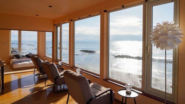 A spacious room with large windows overlooking a snowy landscape. There are two chairs and a small table with a glass. - Nesaki Native (Hakodate)