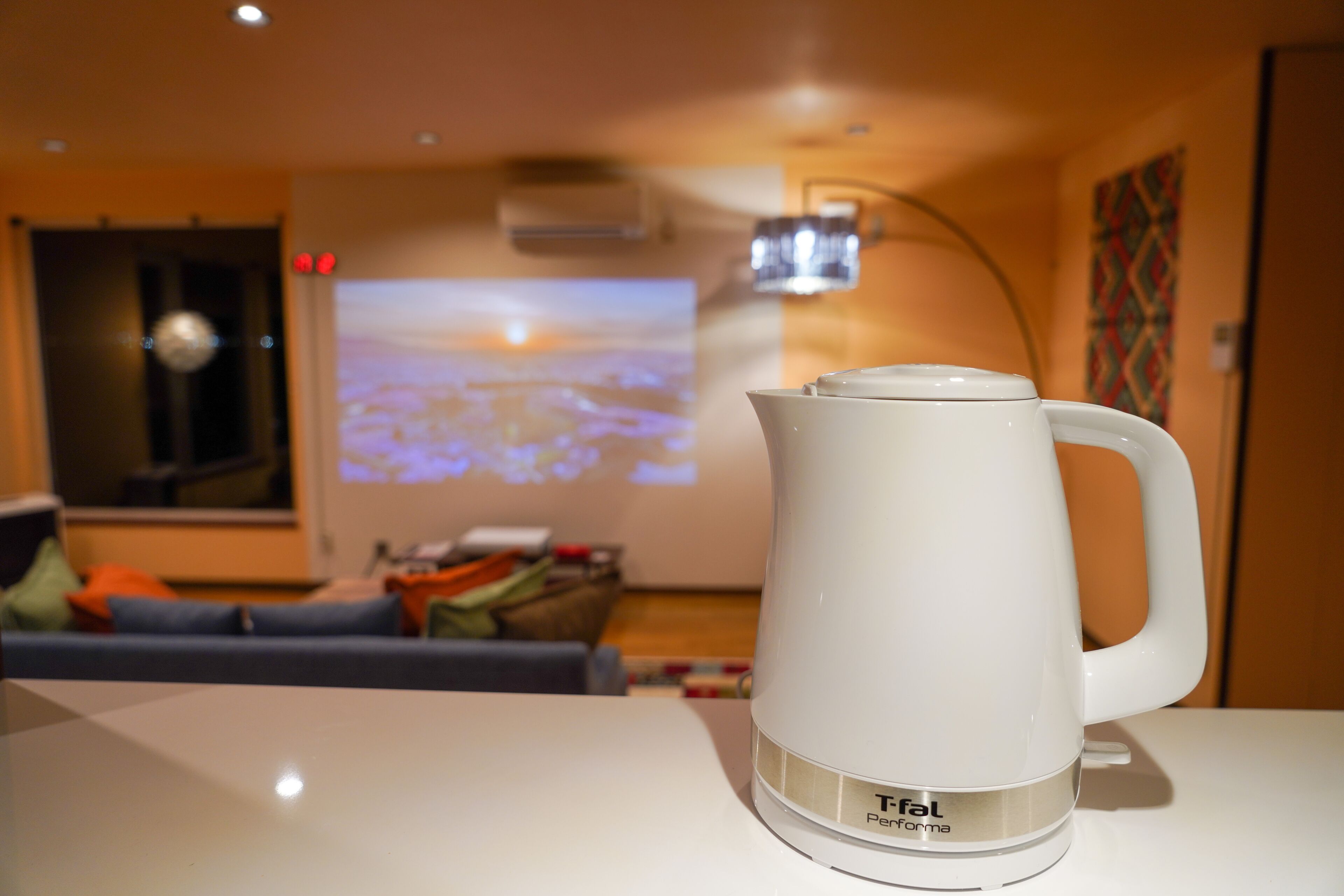 Ein weißes Tefal Performa Wasserkocher auf einem weißen Tisch in einem Raum mit einem blauen Sofa, einem Projektor, der eine Landschaft zeigt, und einem farbenfrohen abstrakten Gemälde an der Wand.