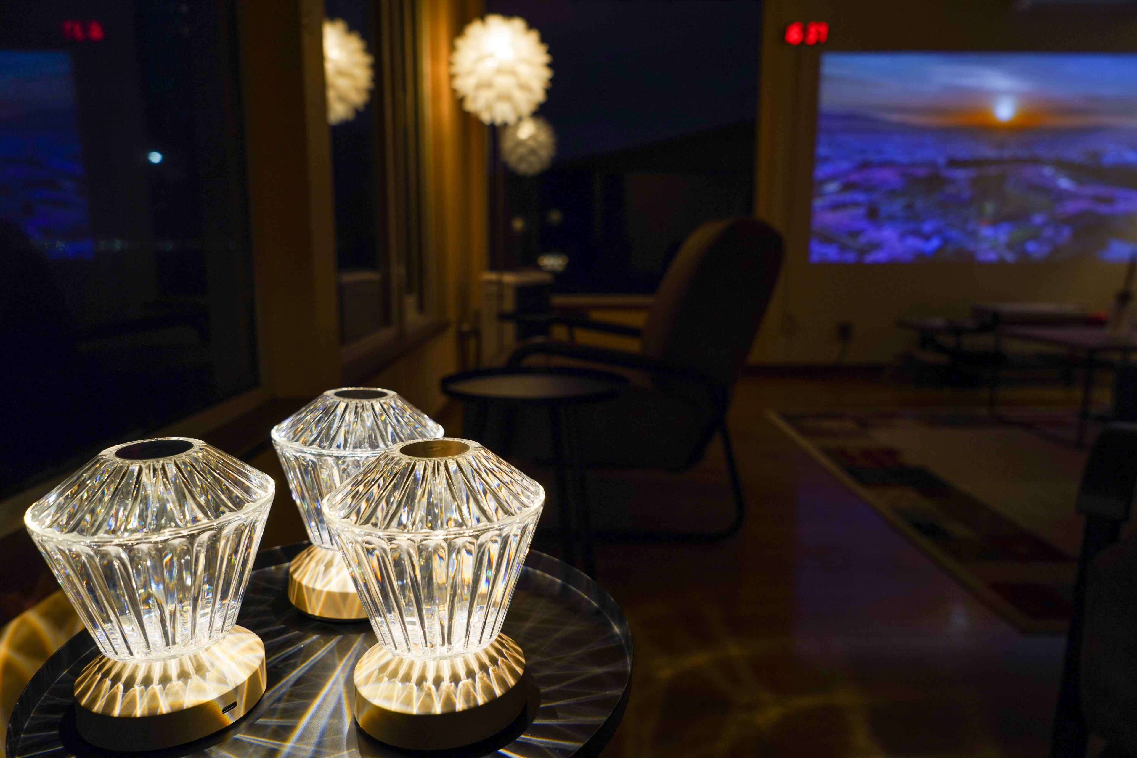A room with a large window, a projection screen displaying a sunset, and three illuminated glass lamps on a table.