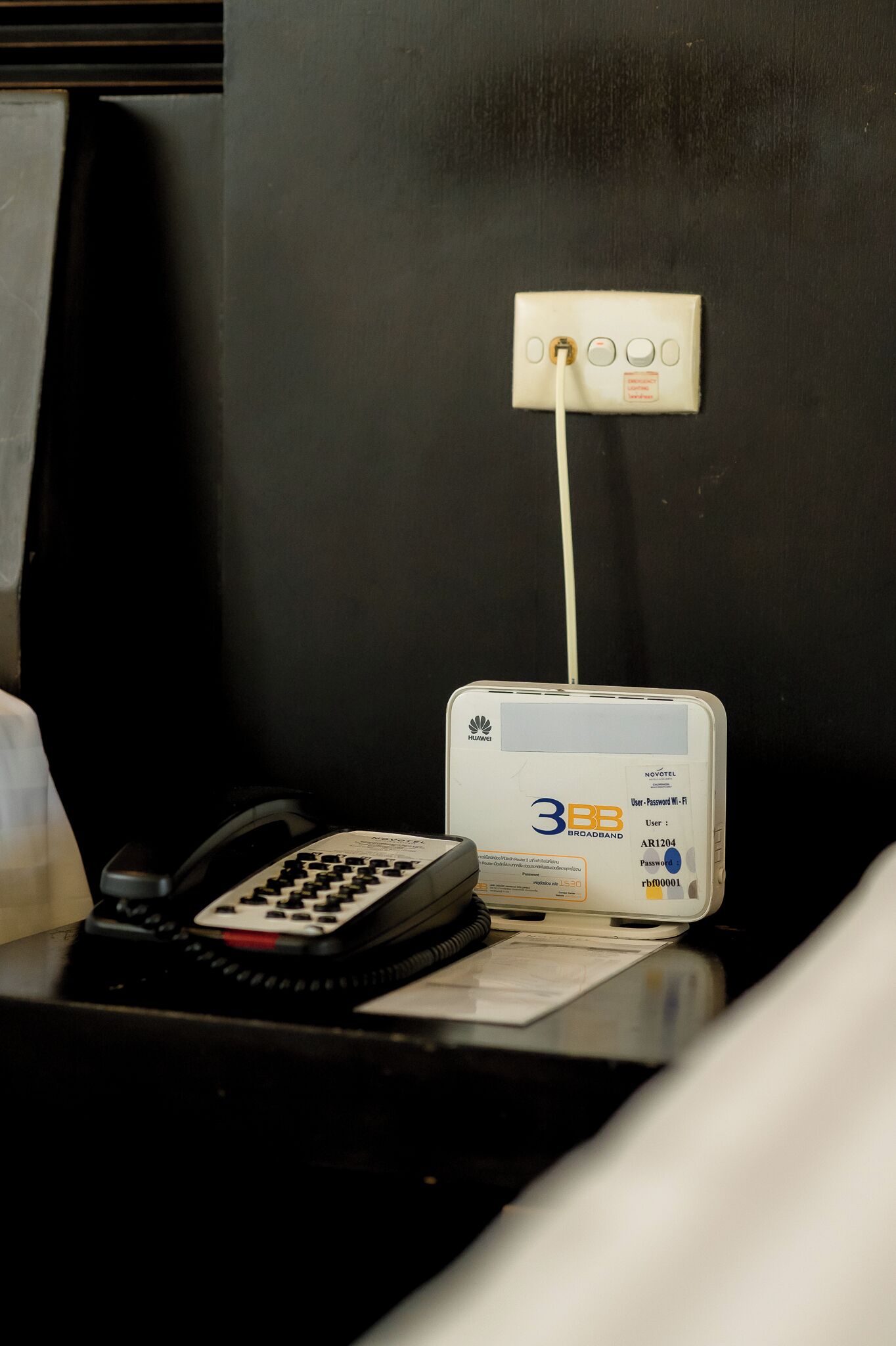 A hotel room with a desk featuring a landline phone, a 3BB broadband router, and a power outlet.