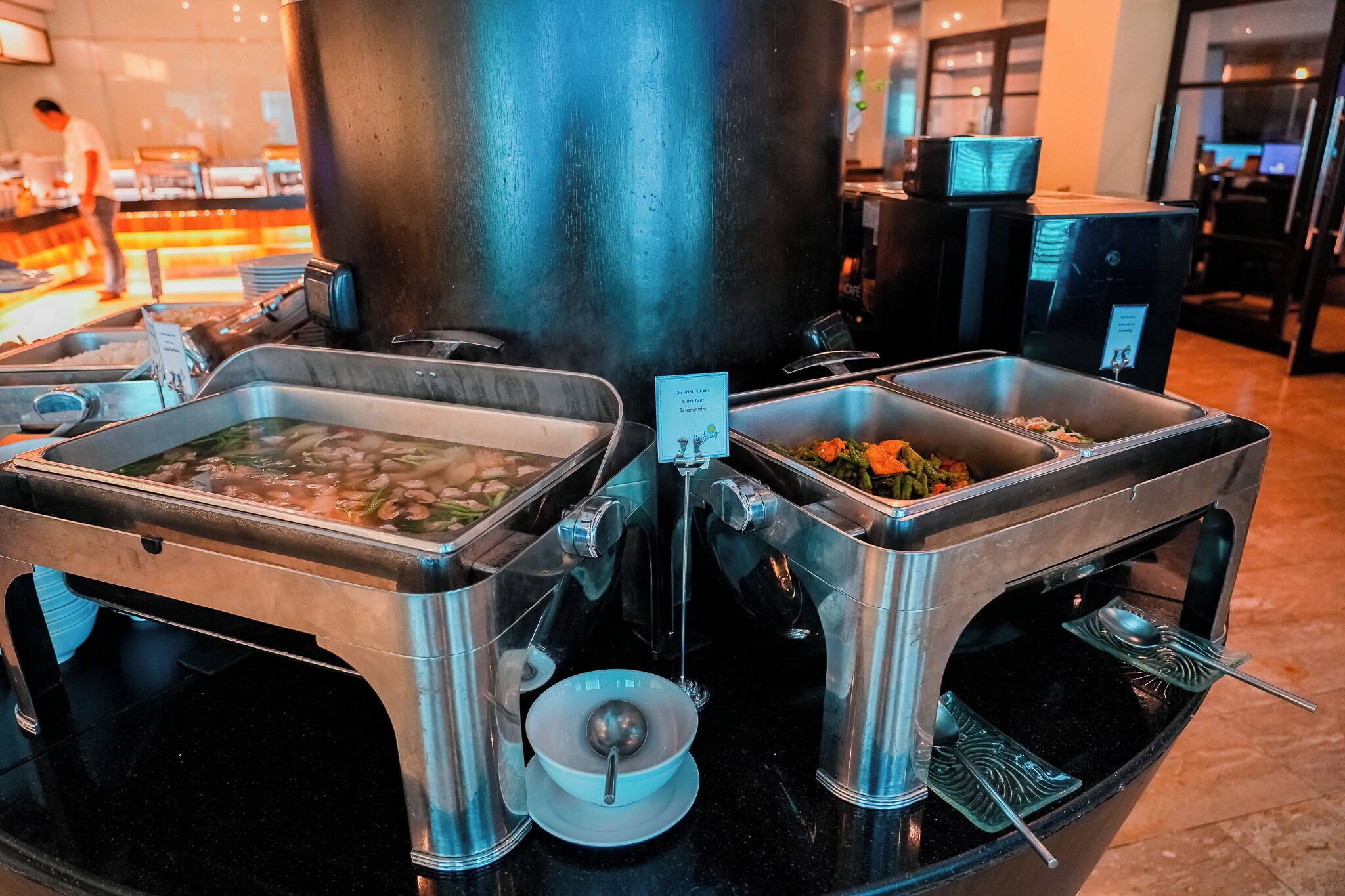 A buffet setup with multiple serving stations, including a hot soup station and a salad station, in a modern hotel setting.