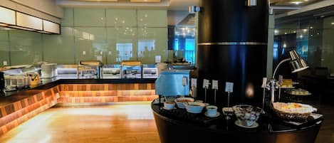 A modern breakfast area with a central coffee station, a seating area with a brick accent wall, and a view of the city through large windows.