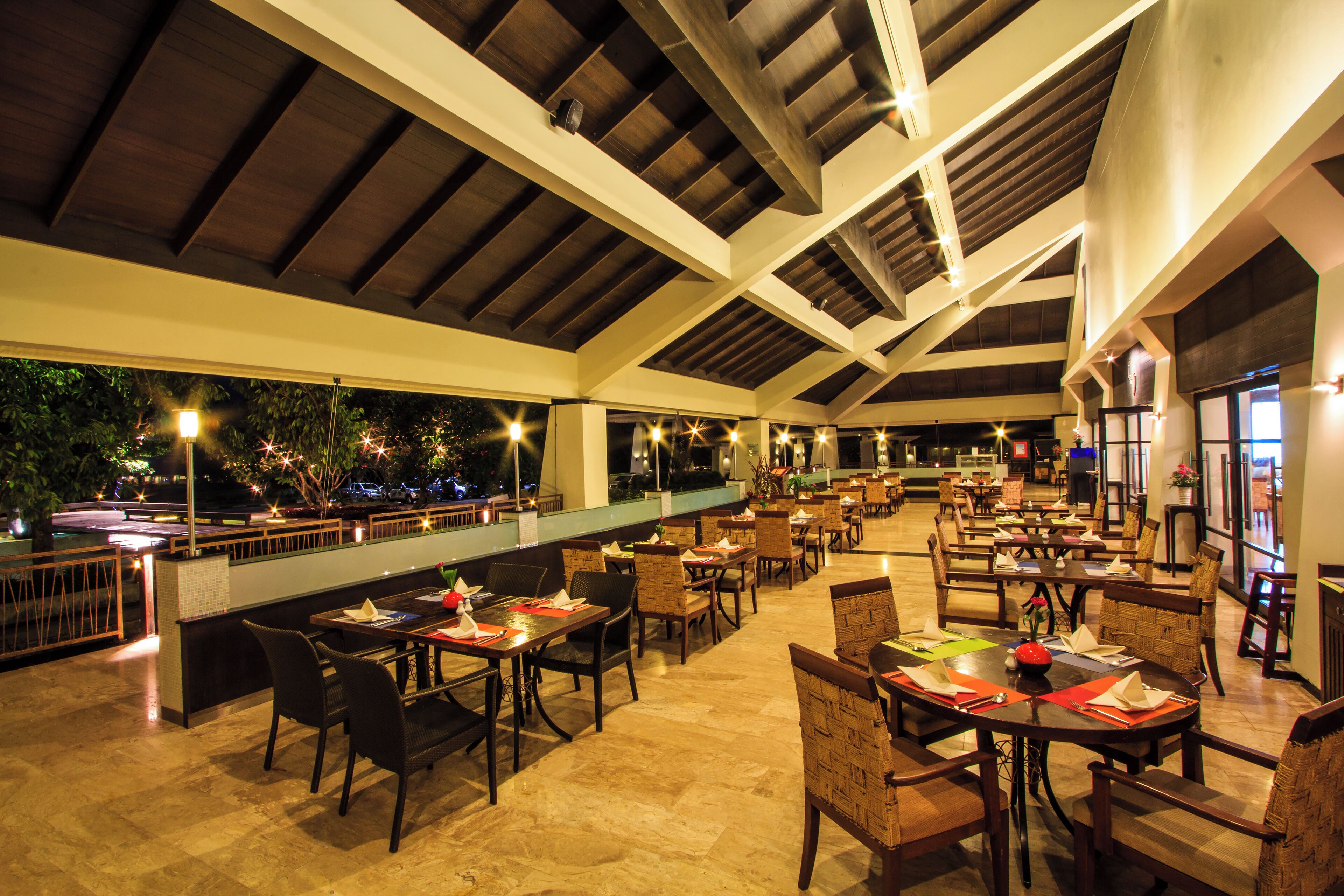 An indoor dining area with multiple tables and chairs, a high ceiling with wooden beams, and a view of the outdoors.