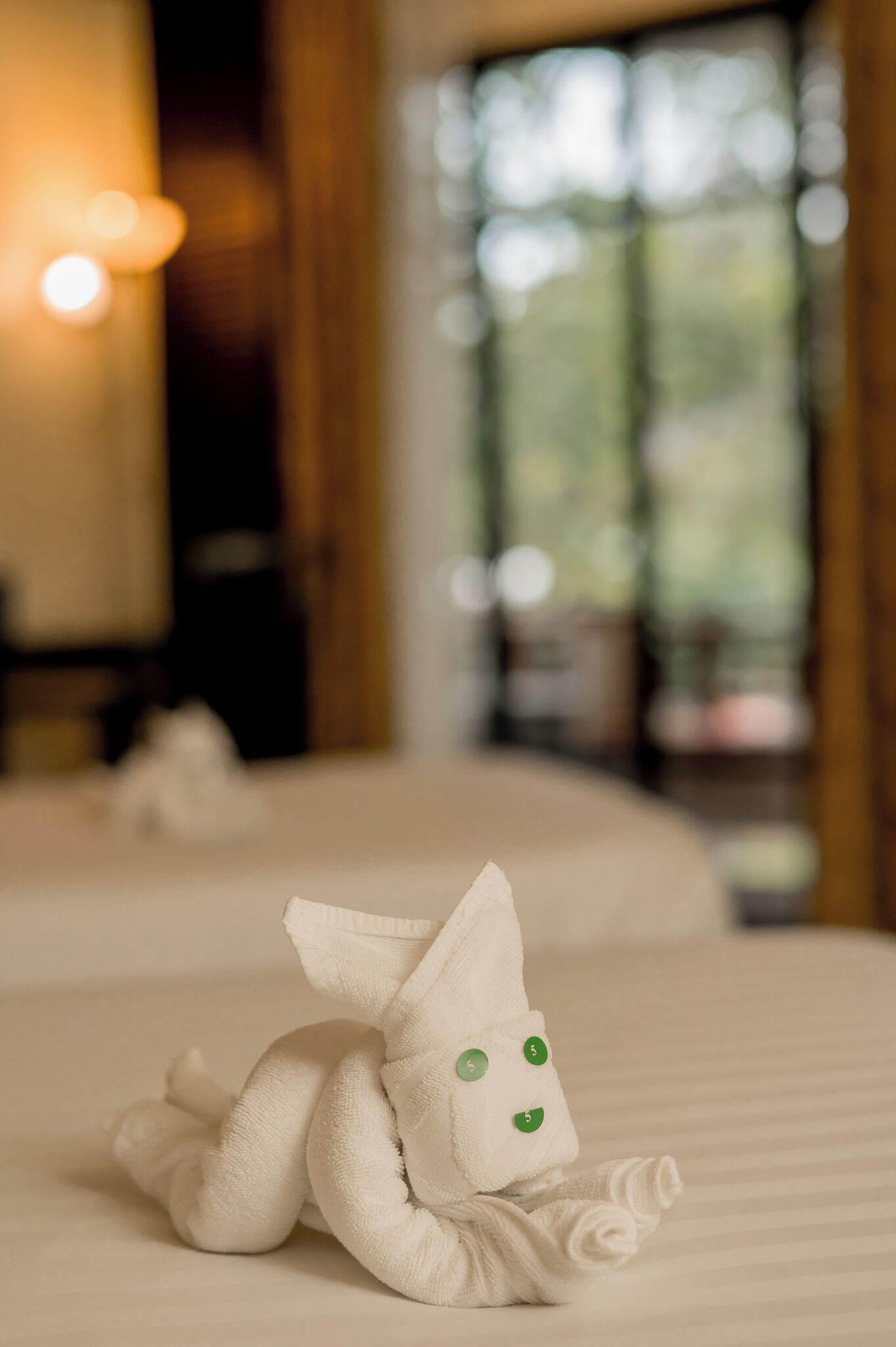 A neatly made towel animal on a bed in a hotel room with a window and a lamp in the background.