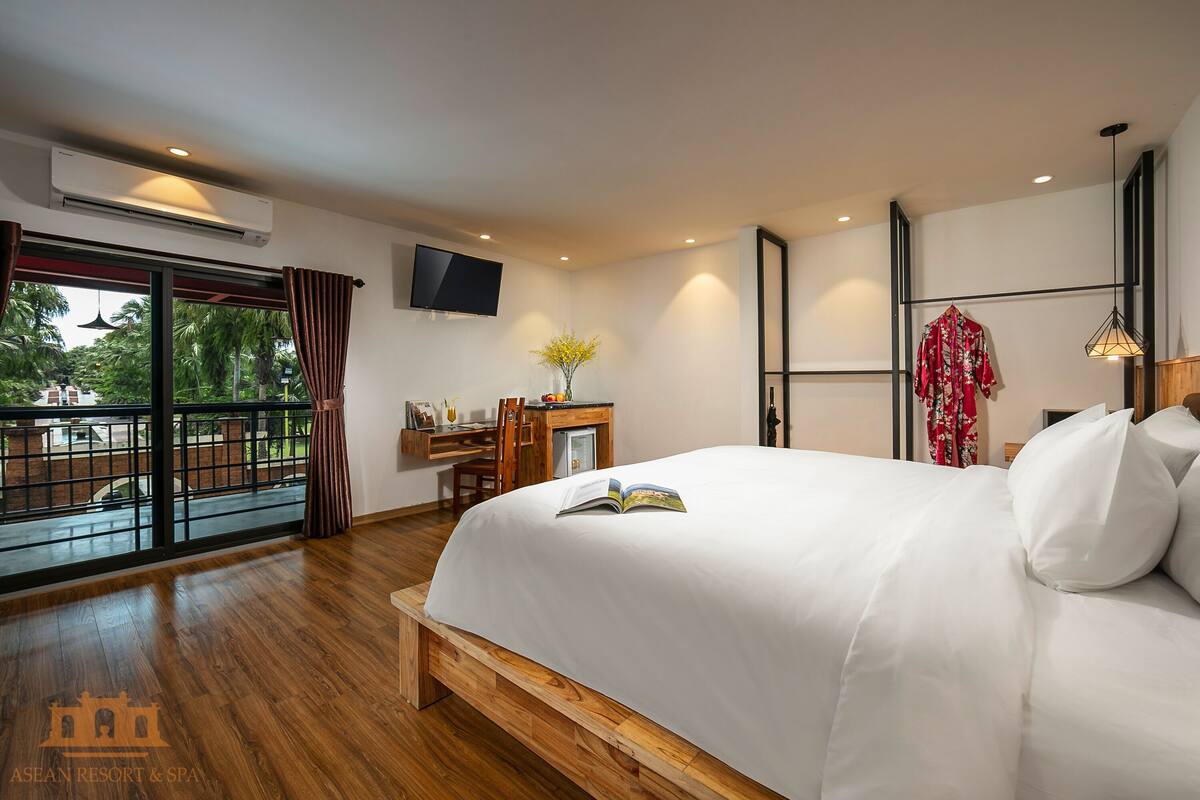 Une chambre d’hôtel moderne dotée d’un grand lit, d’un bureau avec une chaise, d’une télévision et d’un balcon offrant une vue sur la verdure.