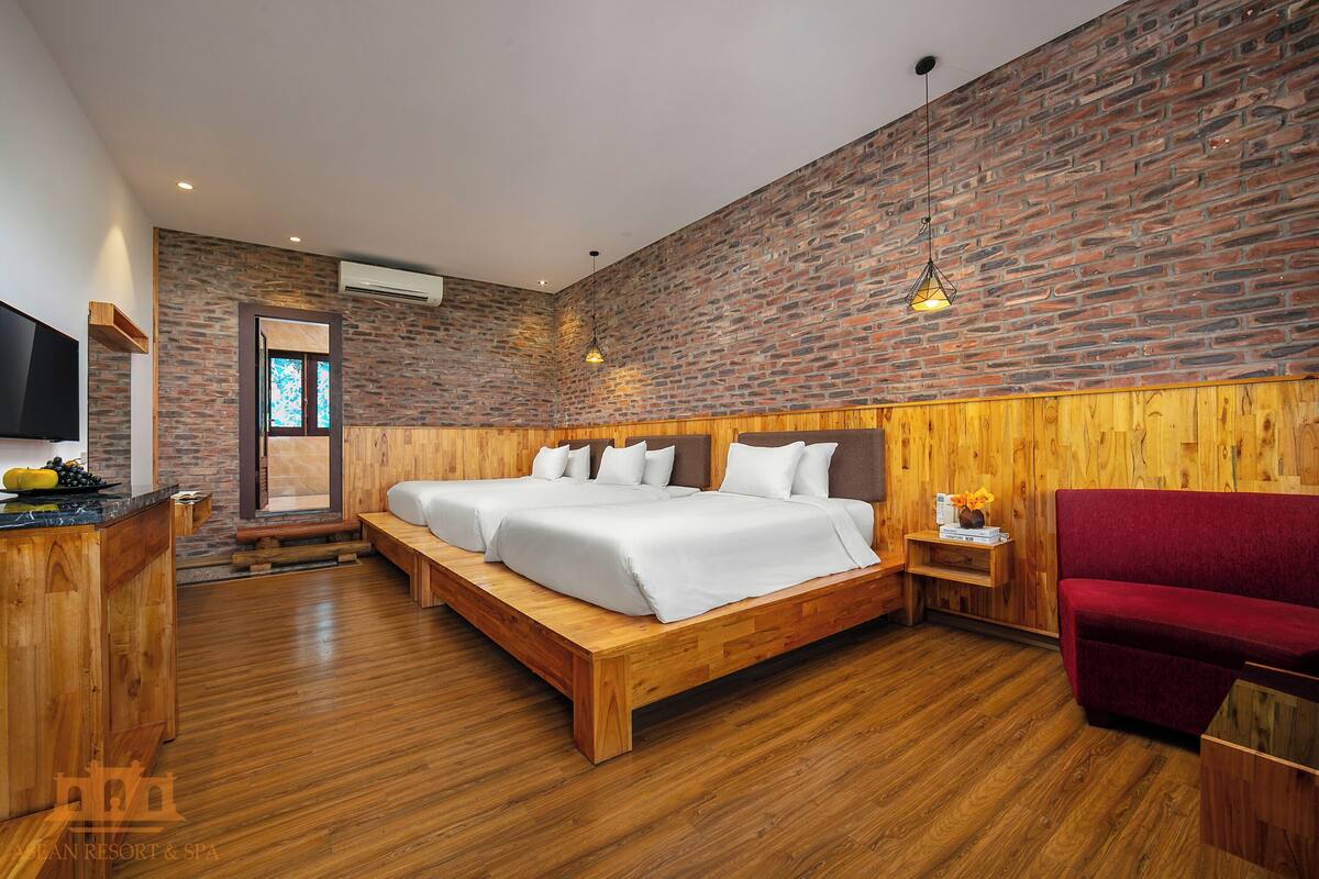 Une chambre d’hôtel moderne avec un mur en briques, un plancher en bois, un grand lit avec du linge de lit blanc, un fauteuil rouge et un banc en bois.